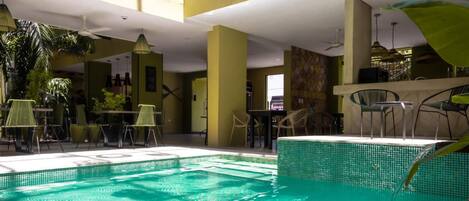 An indoor swimming pool with tiled walls, surrounded by tables and chairs, and a ceiling fan.