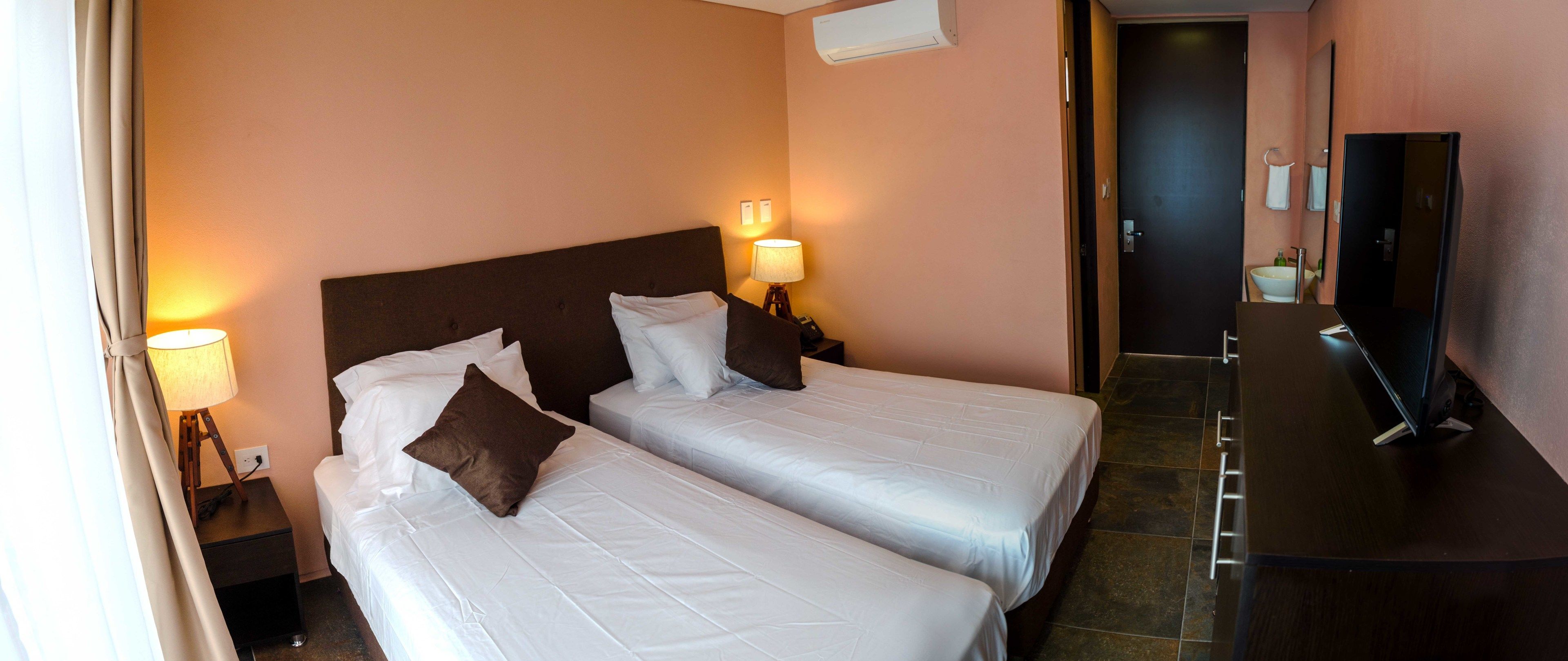 Habitación de hotel con dos camas, una mesita de noche con lámpara, baño visible a través de una puerta abierta y televisor sobre un mueble de madera.