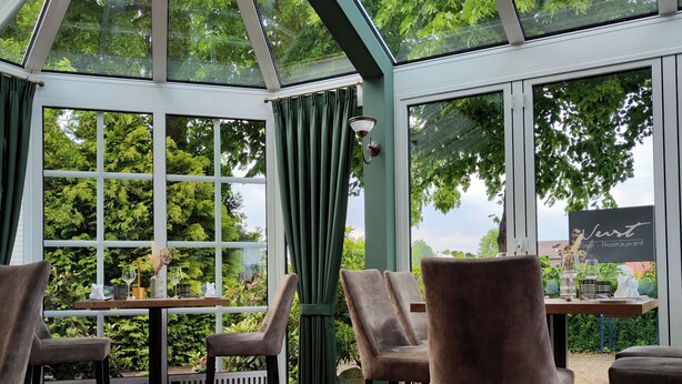 A conservatory with a glass ceiling, wooden flooring, and seating areas with chairs and tables. - Hotel Restaurant Verst (Gronau (Westphalia))