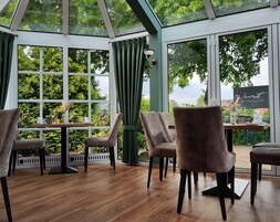 A conservatory with a glass ceiling, wooden flooring, and seating areas with chairs and tables.