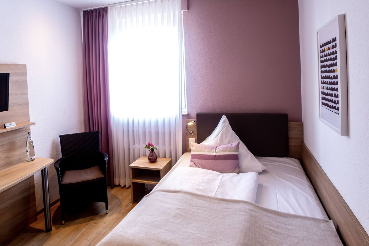A hotel room with a bed, a desk, a chair, a nightstand, a TV, and a window with curtains.