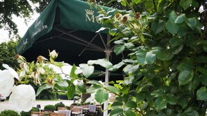 An outdoor seating area with wooden tables and chairs, surrounded by greenery and white flowers. - Hotel Restaurant Verst (Gronau (Westphalia))