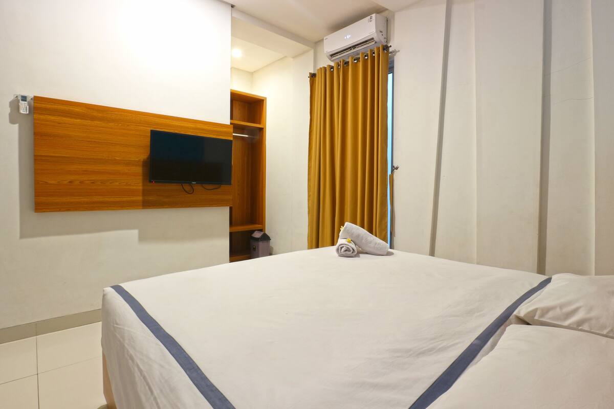A hotel room with a bed, a mounted television, an air conditioning unit, and a curtain.