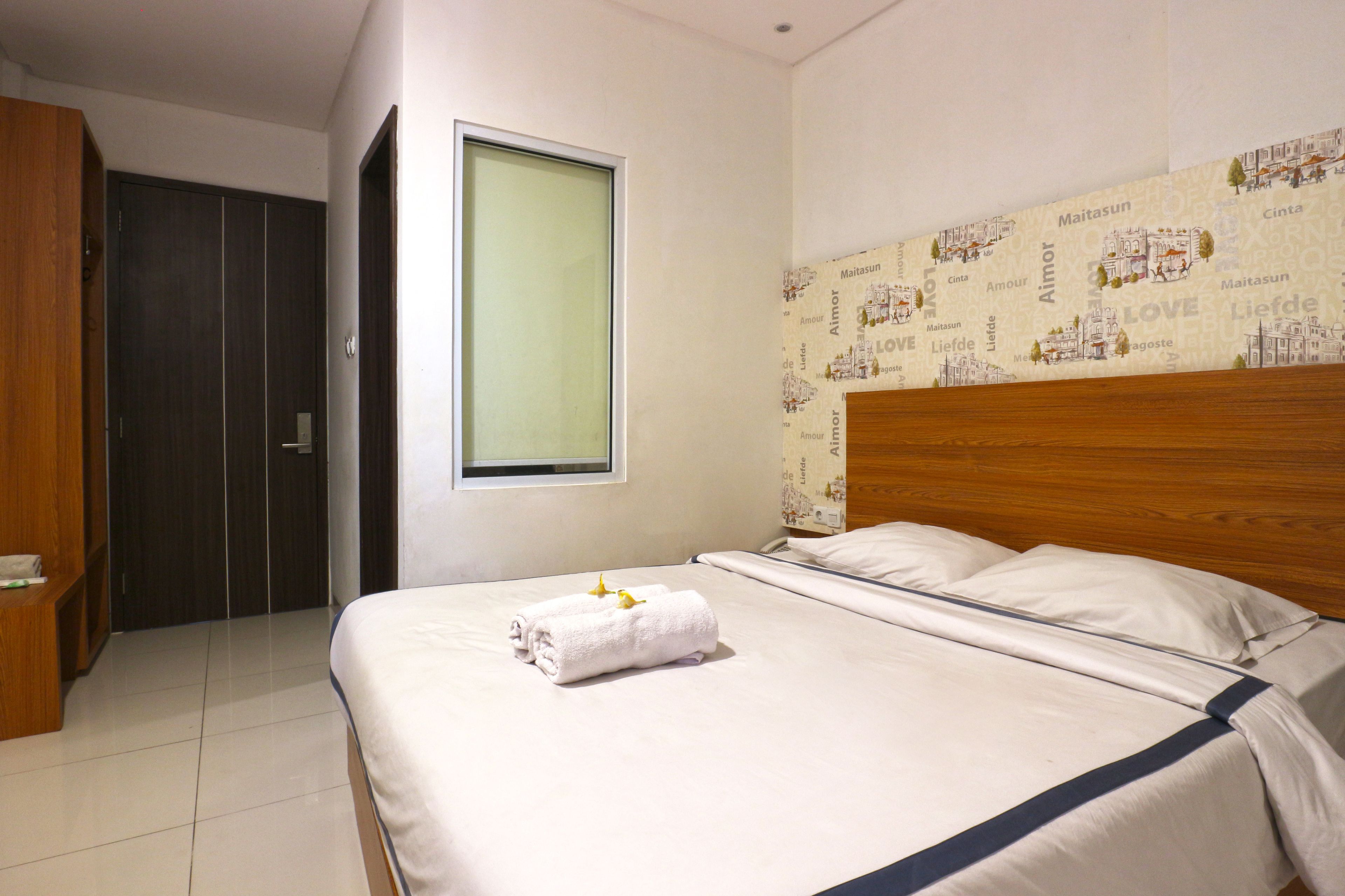 A neatly made bed with a blue accent, a wooden wardrobe, and a window with a view of a cityscape.