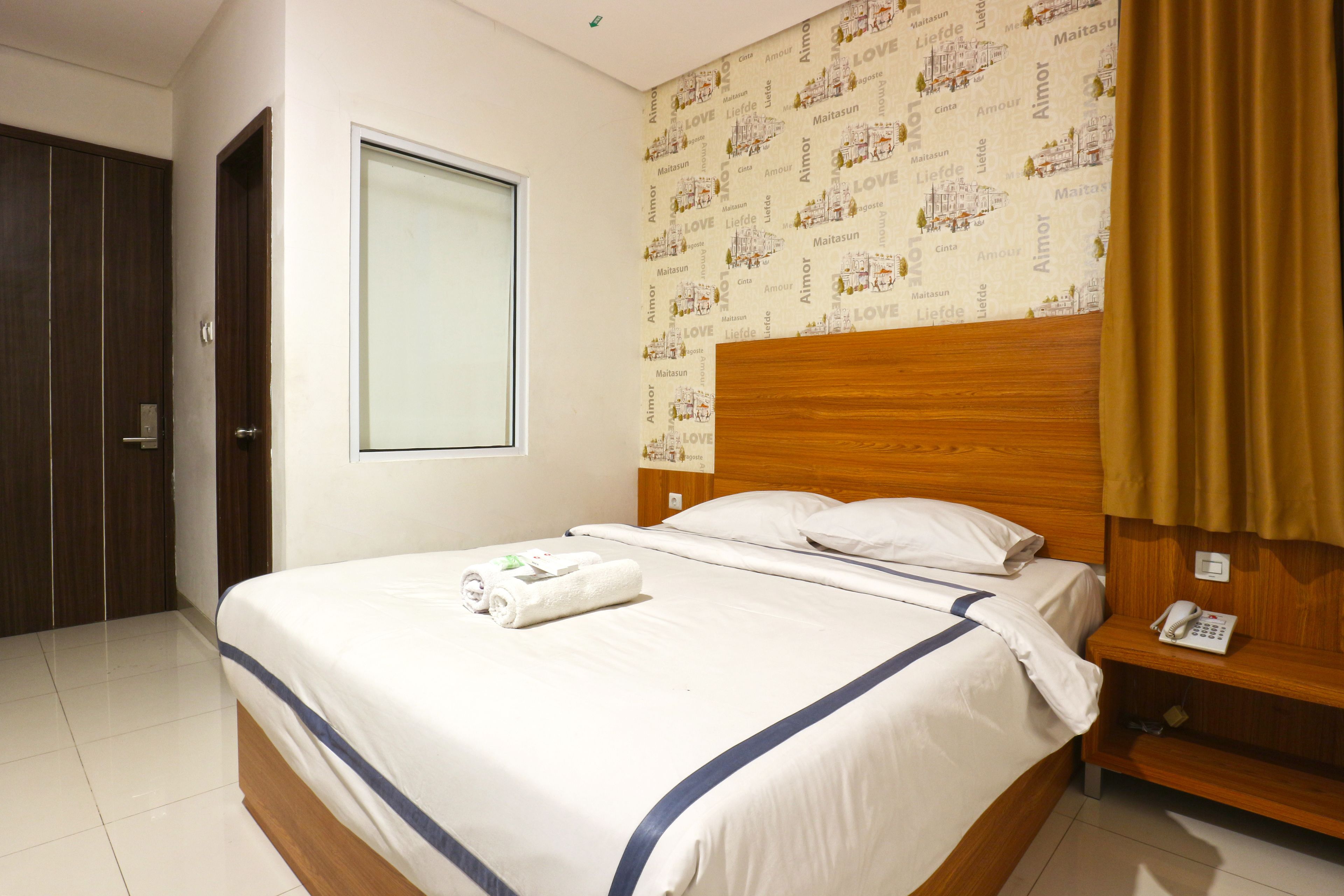 A hotel room with a neatly made bed, a wooden headboard, a bedside telephone, and a window with a curtain.