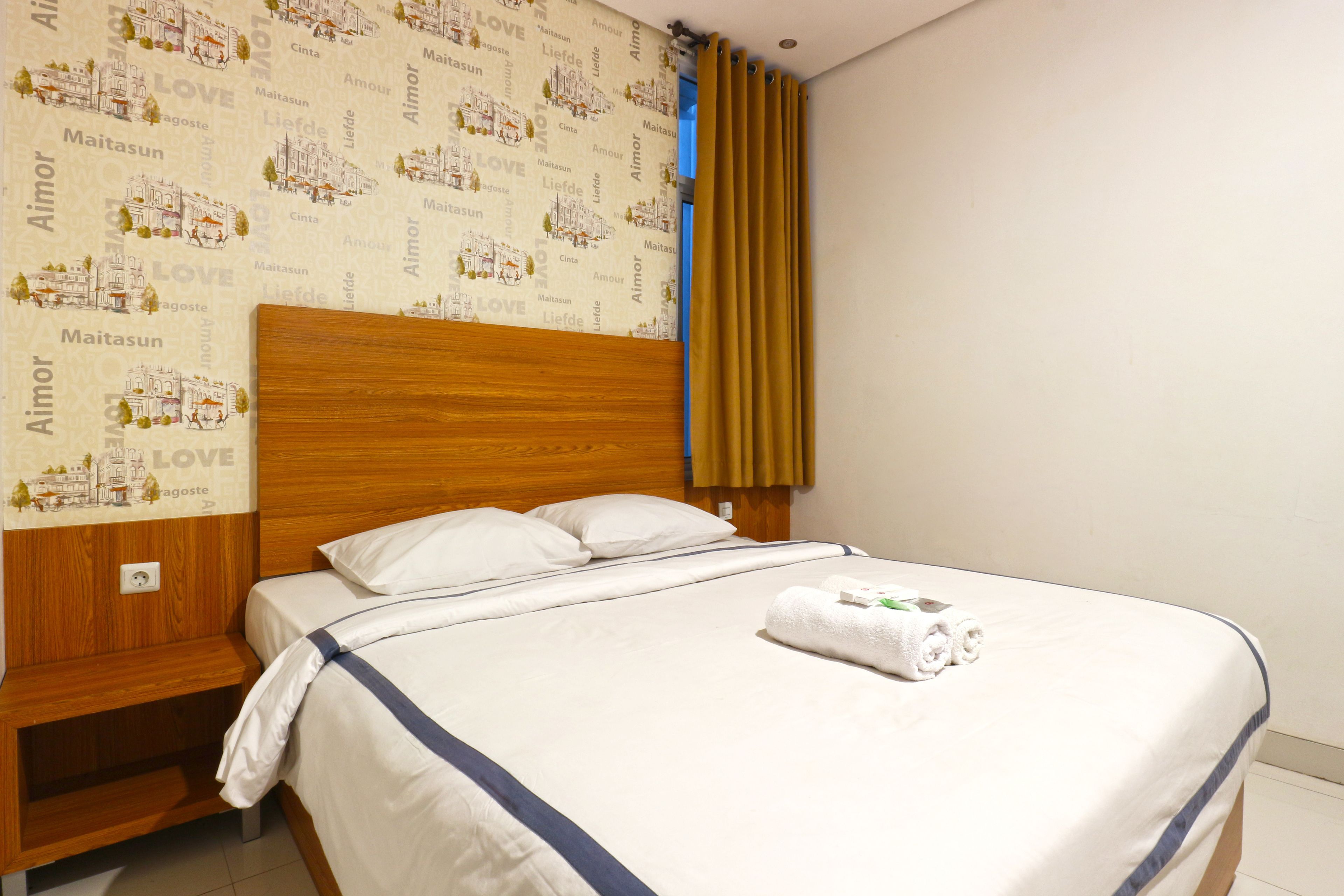A neatly made bed with white linens and a blue accent, a wooden headboard, and a wall with a repeating pattern of 'AIMOR' and 'LOVE'.