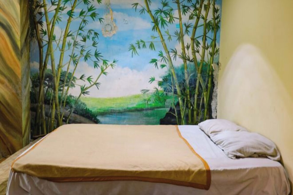 A bed with a beige and white bedspread in a room with a mural of bamboo and a river.