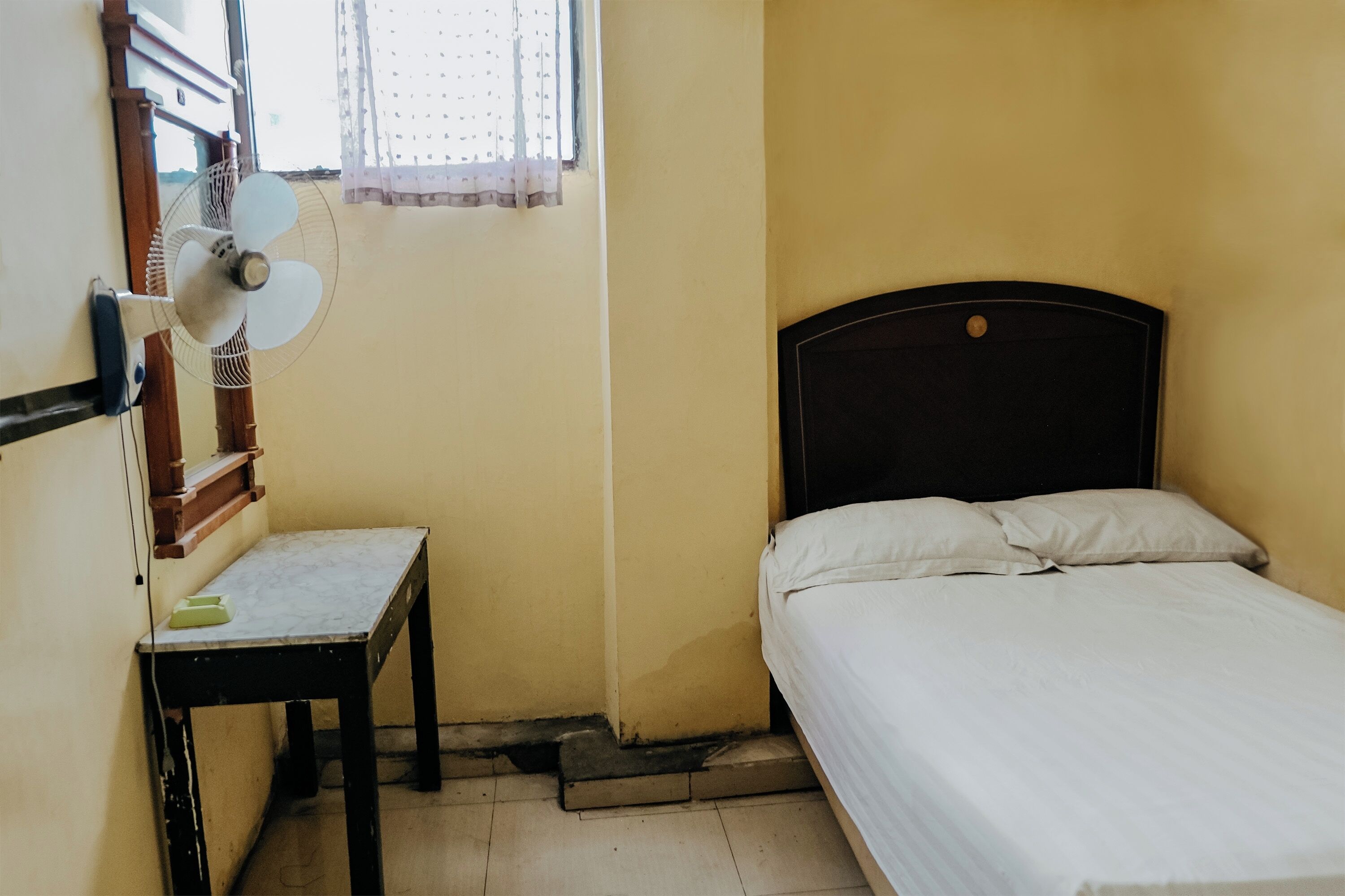 A small room with a bed, a desk, and a standing fan.