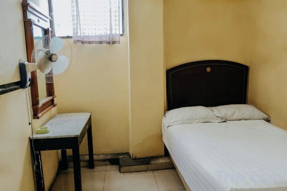 A small room with a bed, a table, and a standing fan.