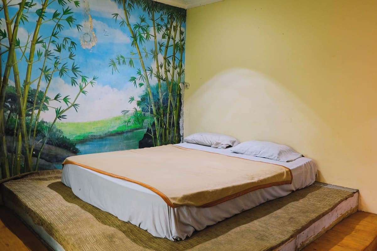 A single bed with a white and brown bedspread, a wooden platform, and a mural of bamboo and a landscape on the wall.