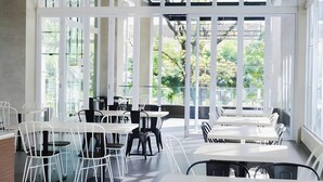 A modern dining area with white tables and chairs, large windows, and a view of greenery outside. - Kampioen Bed and Breakfast (Bandung)