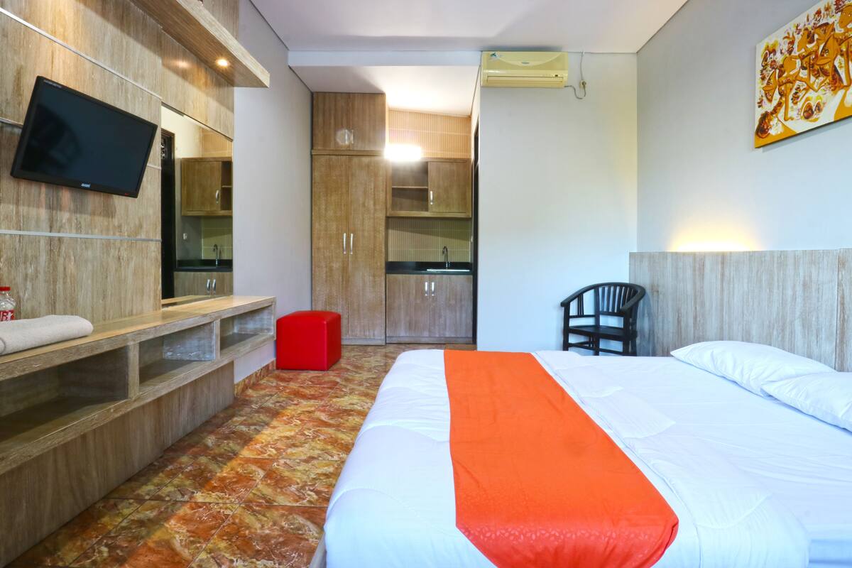 Una habitación de hotel con una cama, un televisor montado en la pared, un taburete rojo y un armario de madera.