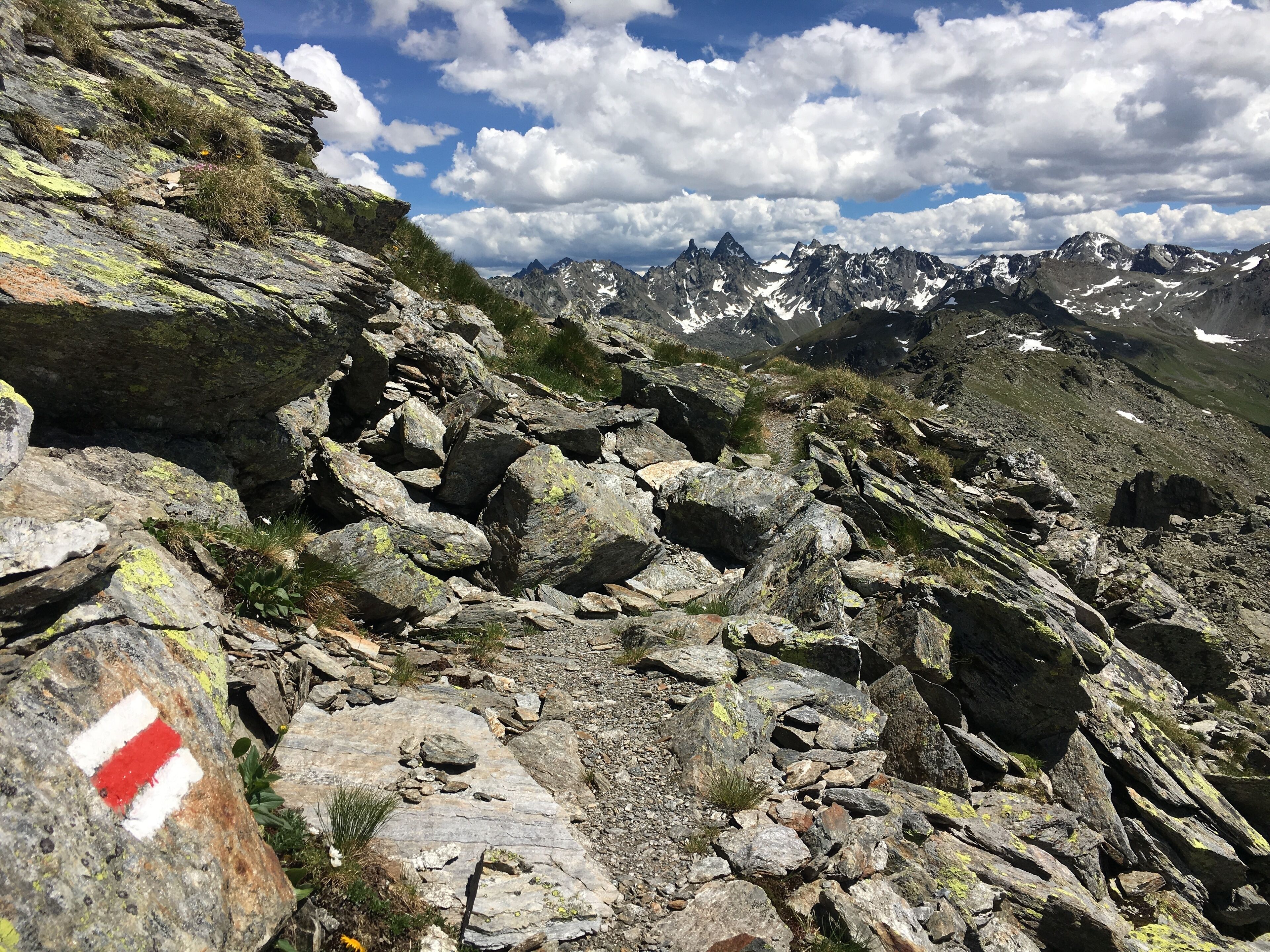 Un sentier de montagne rocailleux, balisé de marques rouges et blanches, serpentant à travers un paysage accidenté et offrant une vue sur des sommets enneigés au loin, sous un ciel partiellement nuageux.