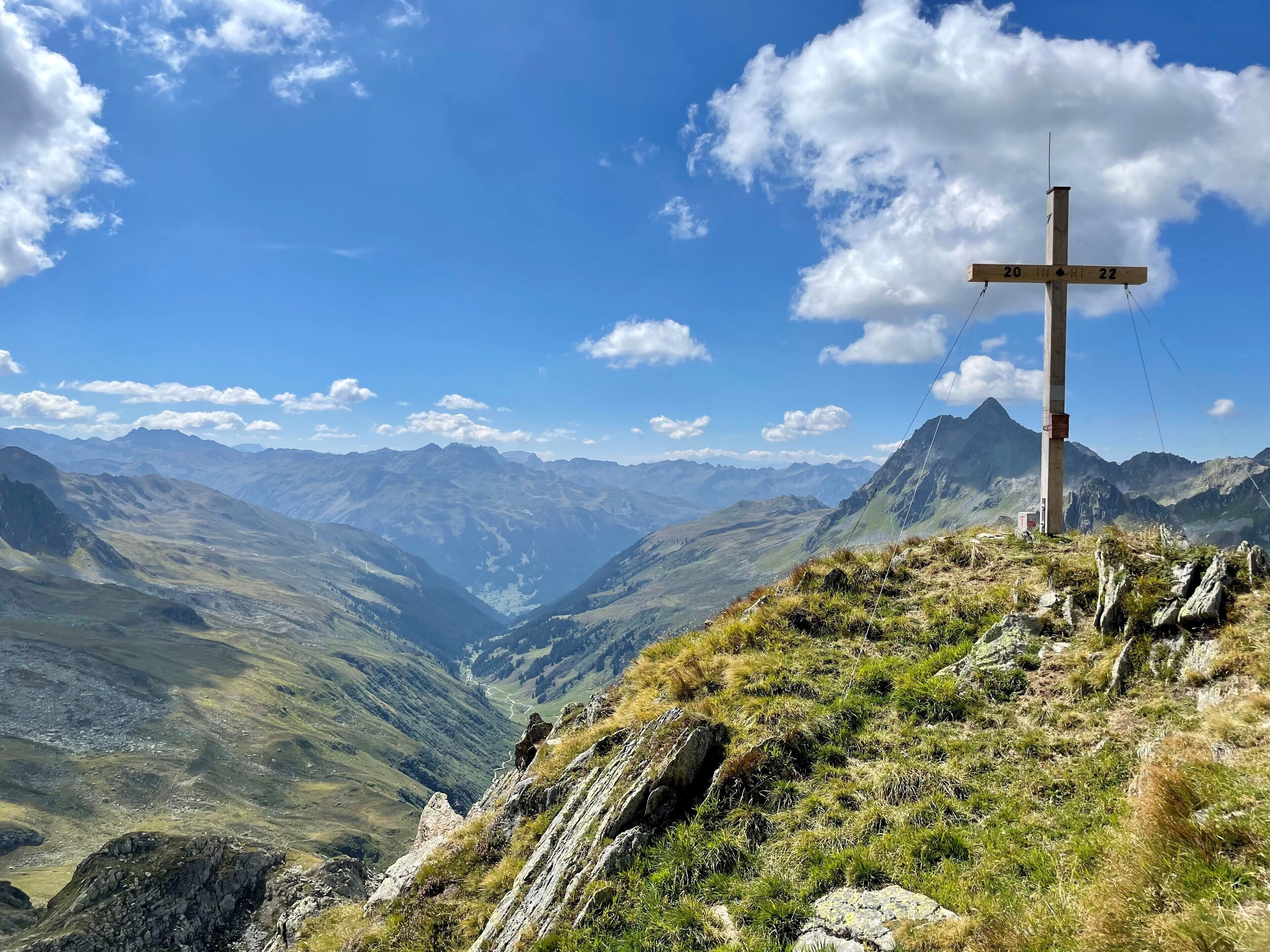 A wooden cross on a mountain peak with a valley and distant mountains in the background.