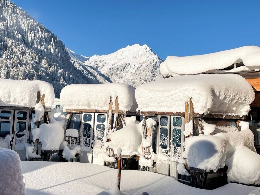 Una fila de autobuses cubiertos de nieve estacionados en una zona nevada, con montañas al fondo.