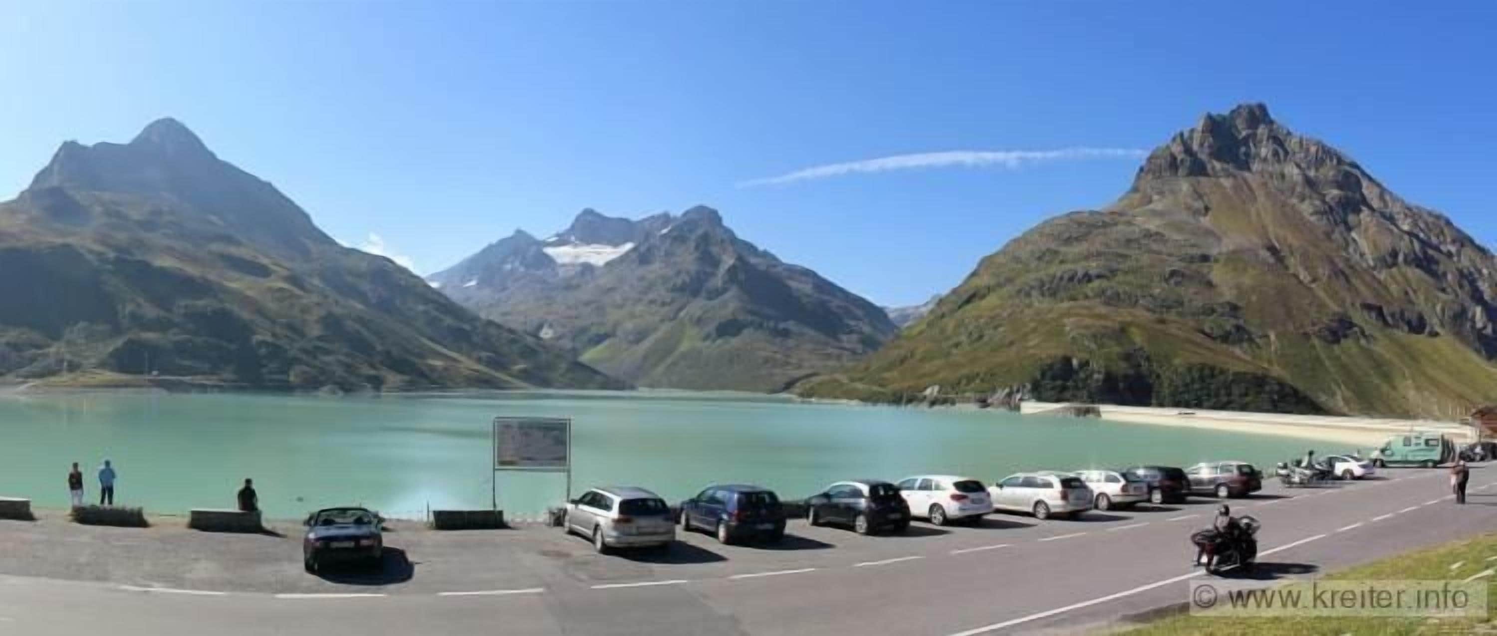 Un área de estacionamiento con vehículos y vistas a montañas y un lago.