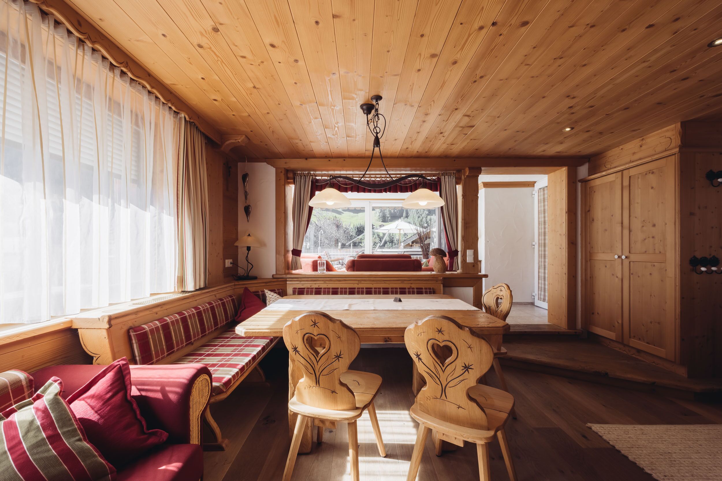 Ein gemütliches Zimmer mit Holzböden und -wänden, einem großen Fenster mit Vorhängen, einer Holzbank und zwei Stühlen mit Herzmotiv.