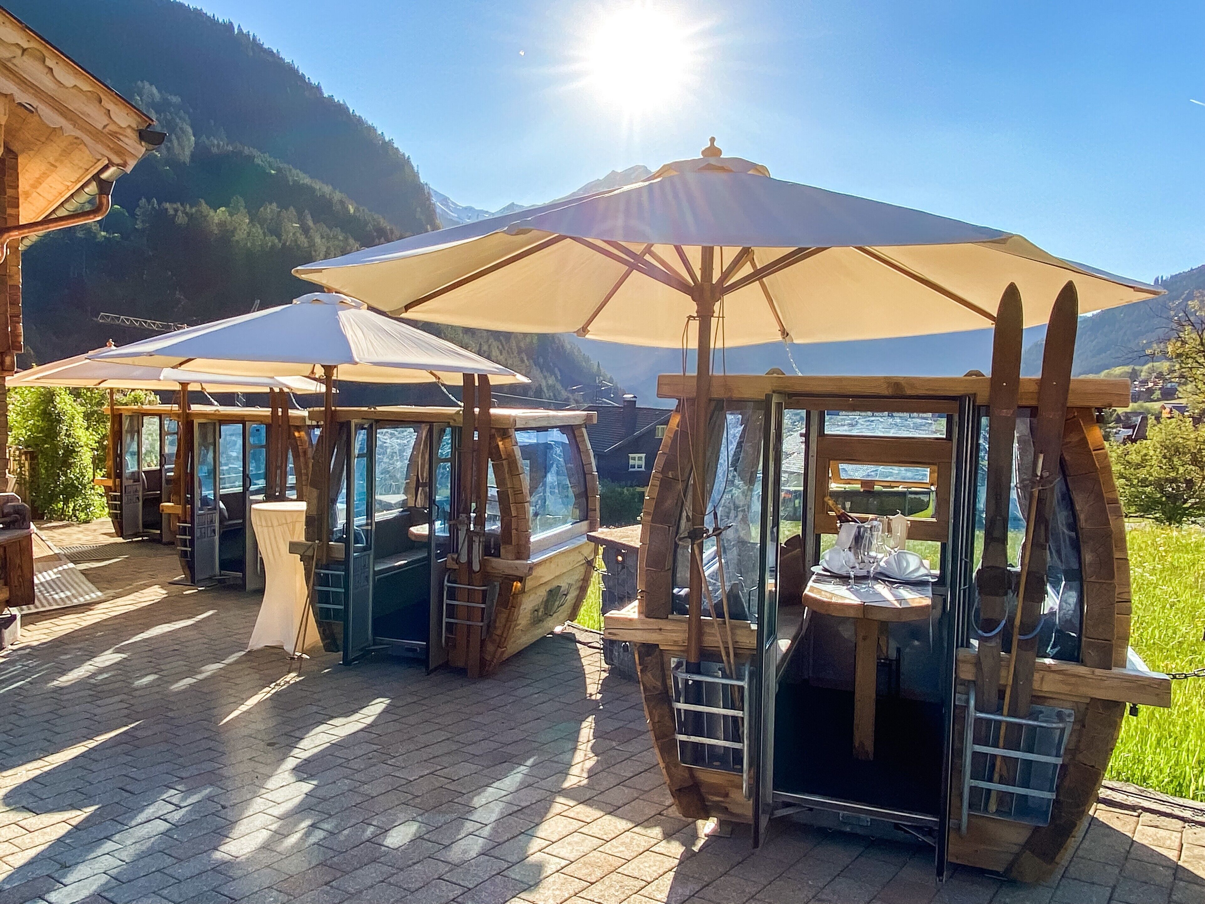 Outdoor wooden structures with glass walls, tables, and chairs, set against a mountainous backdrop.