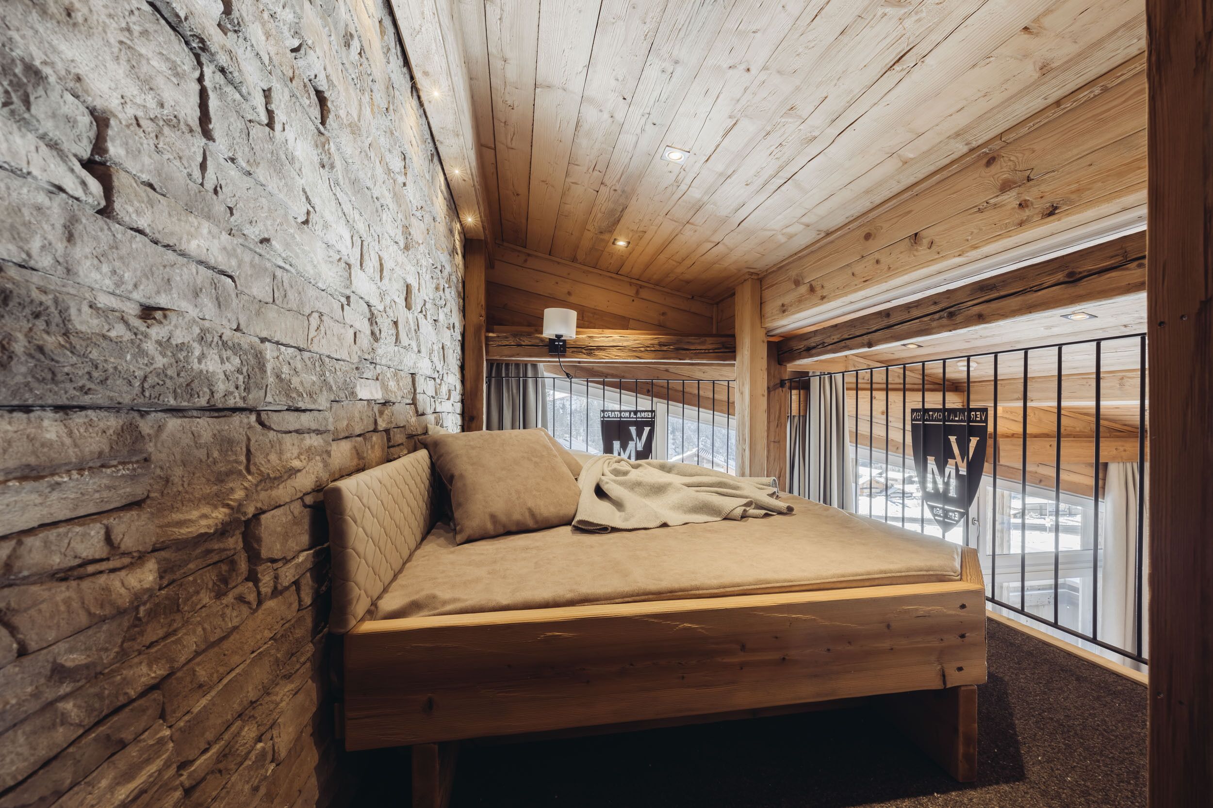 A wooden bed in a cabin with a stone wall, a window with a metal railing, and a ceiling light.