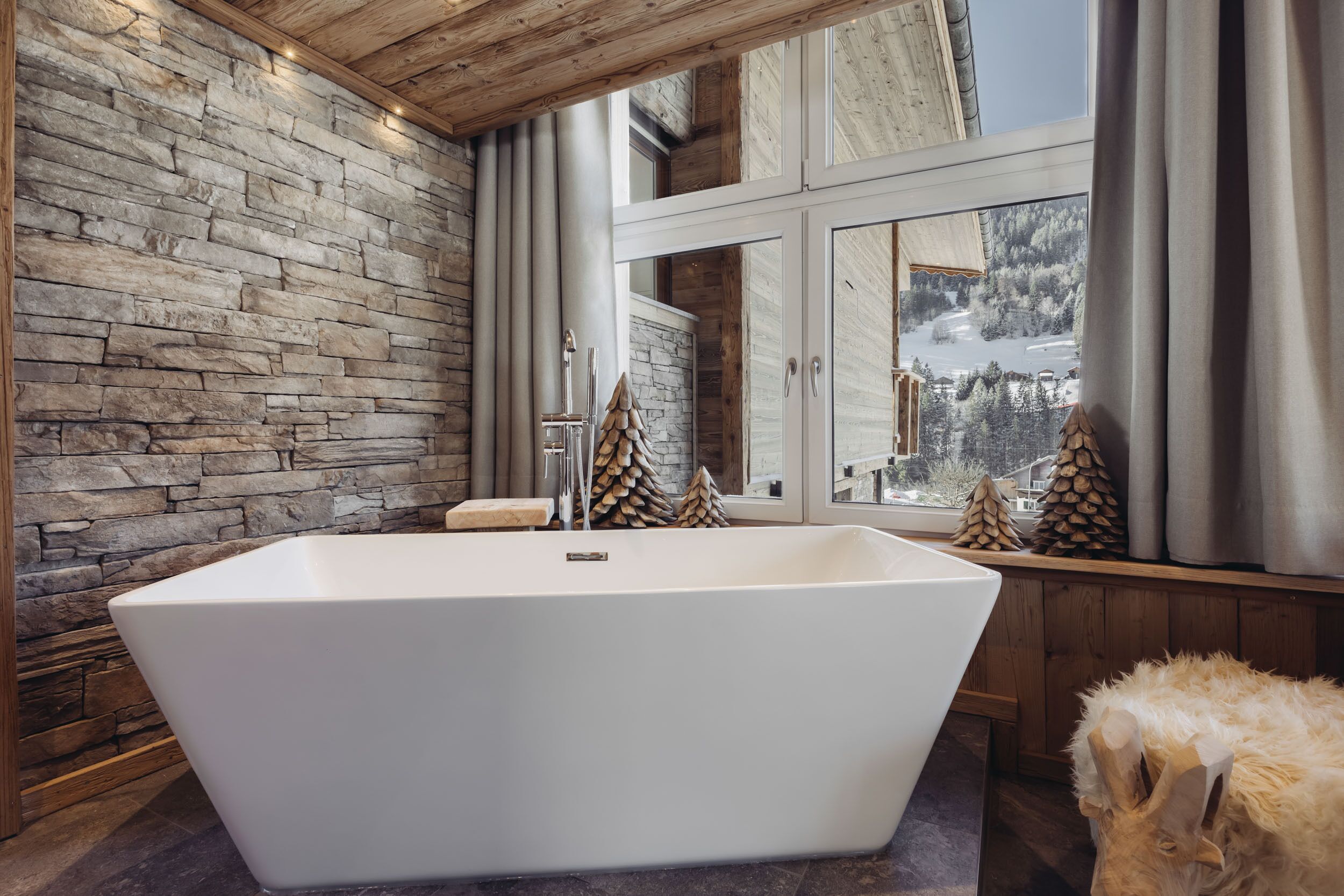 Un bagno moderno con una vasca da bagno indipendente di grandi dimensioni, pareti in pietra e vista su un paesaggio innevato attraverso ampie finestre.