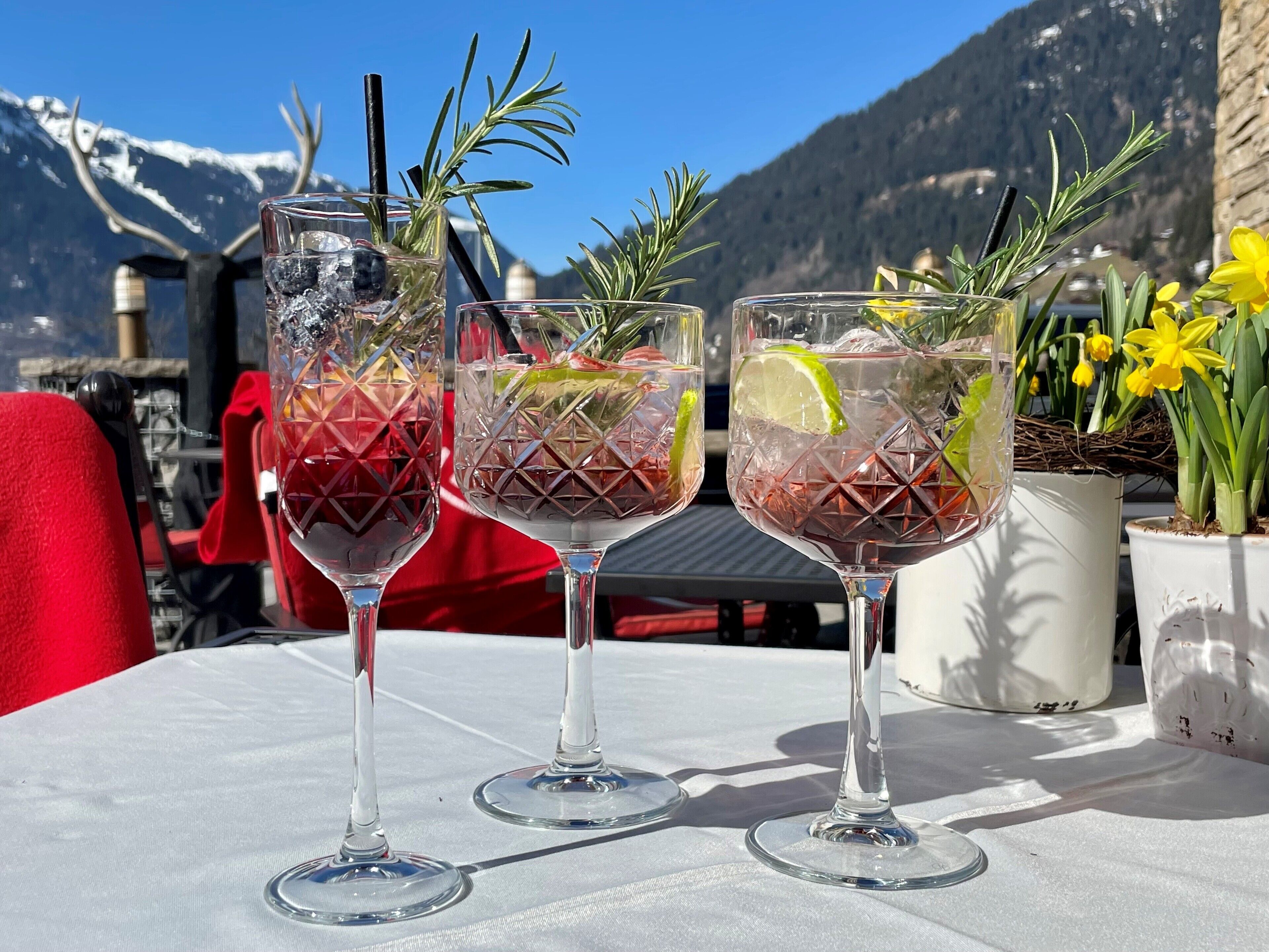 Three glasses of drink with garnishes on a table with a mountain view in the background.