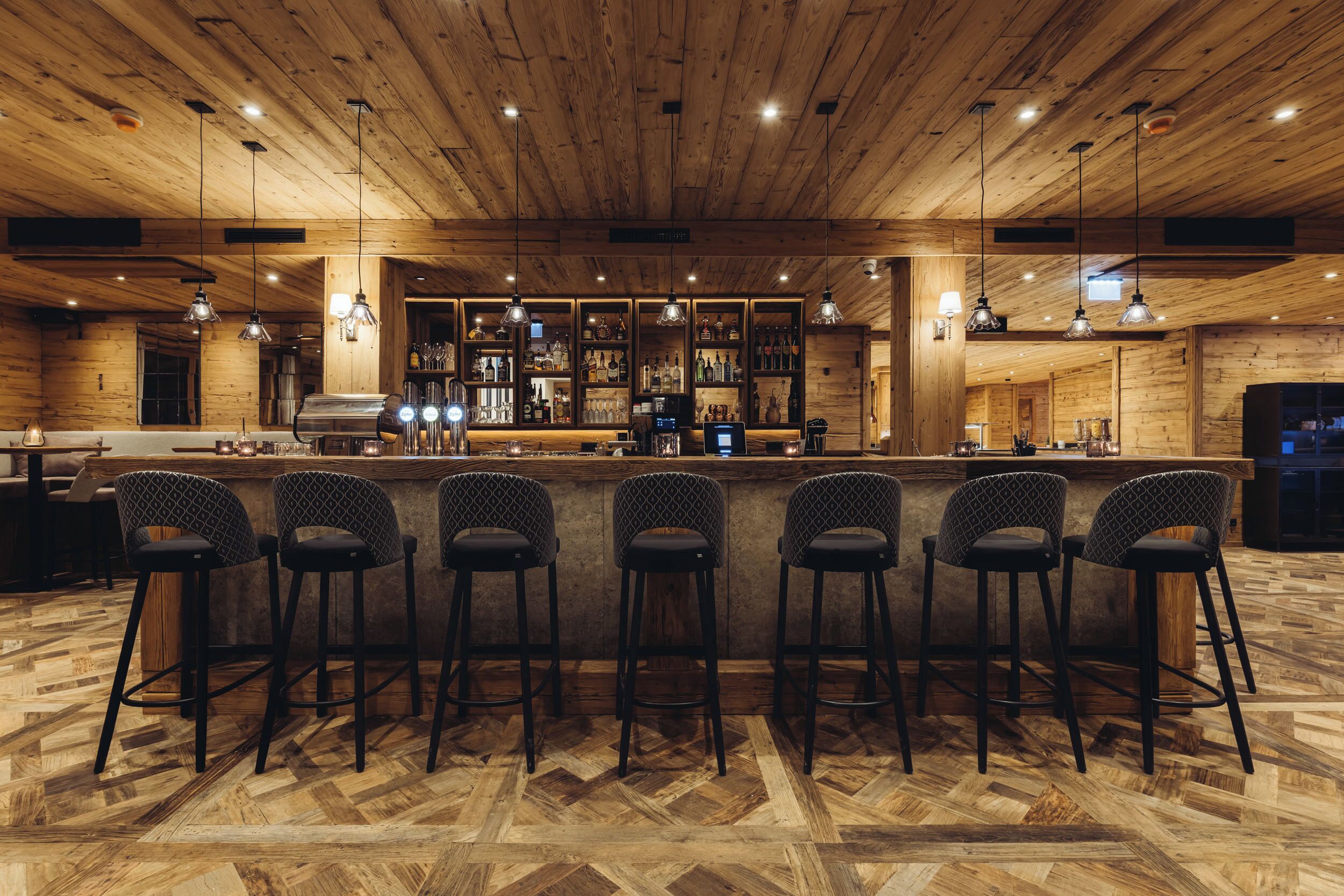 A bar area with wooden ceiling and herringbone floor pattern, bar stools, and shelves with bottles.