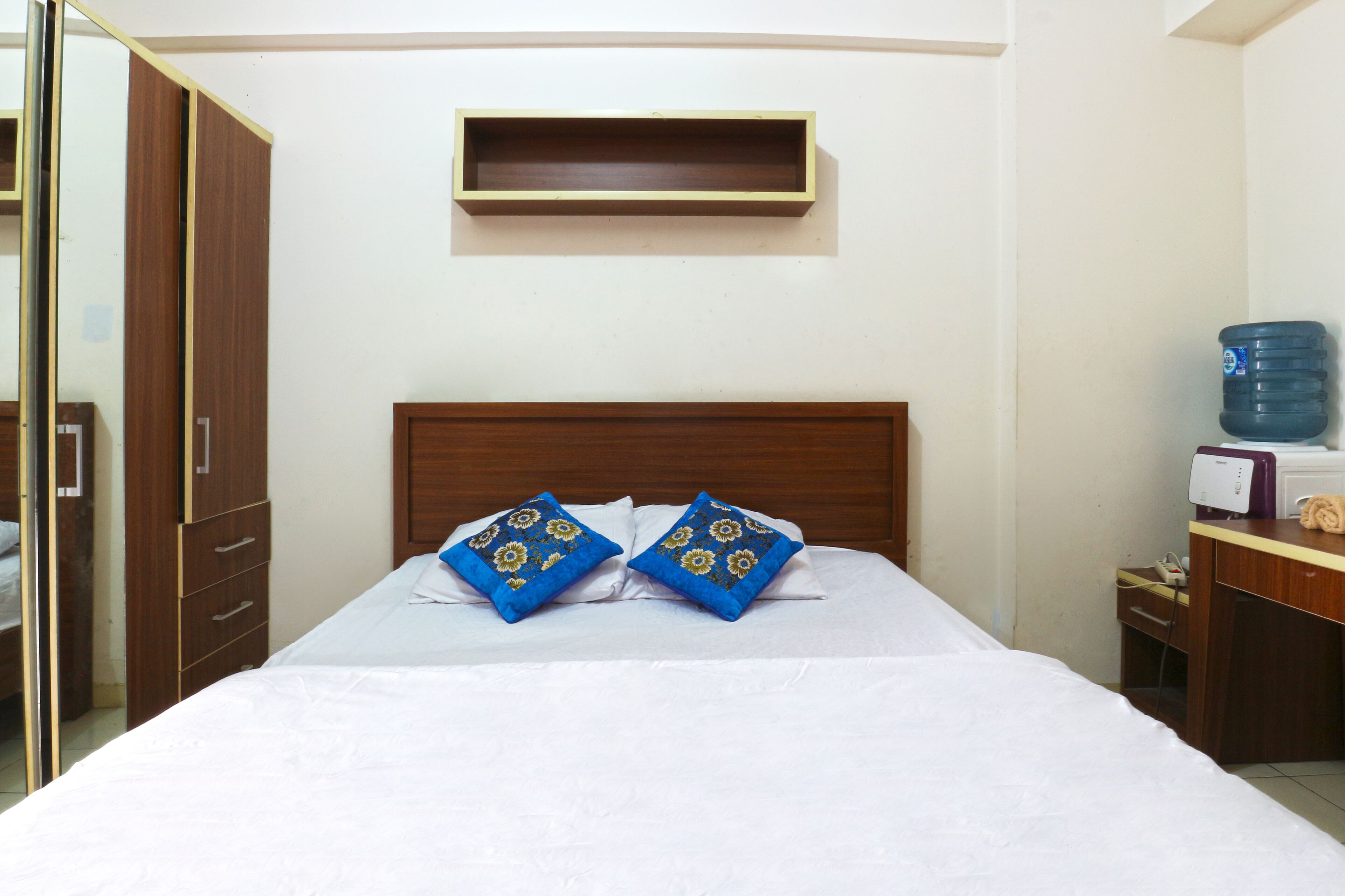 Camera d'albergo con un letto, due cuscini, una testiera in legno, un piccolo ripiano sopra il letto, un dispenser d'acqua e una scrivania con un telefono e una lampada.