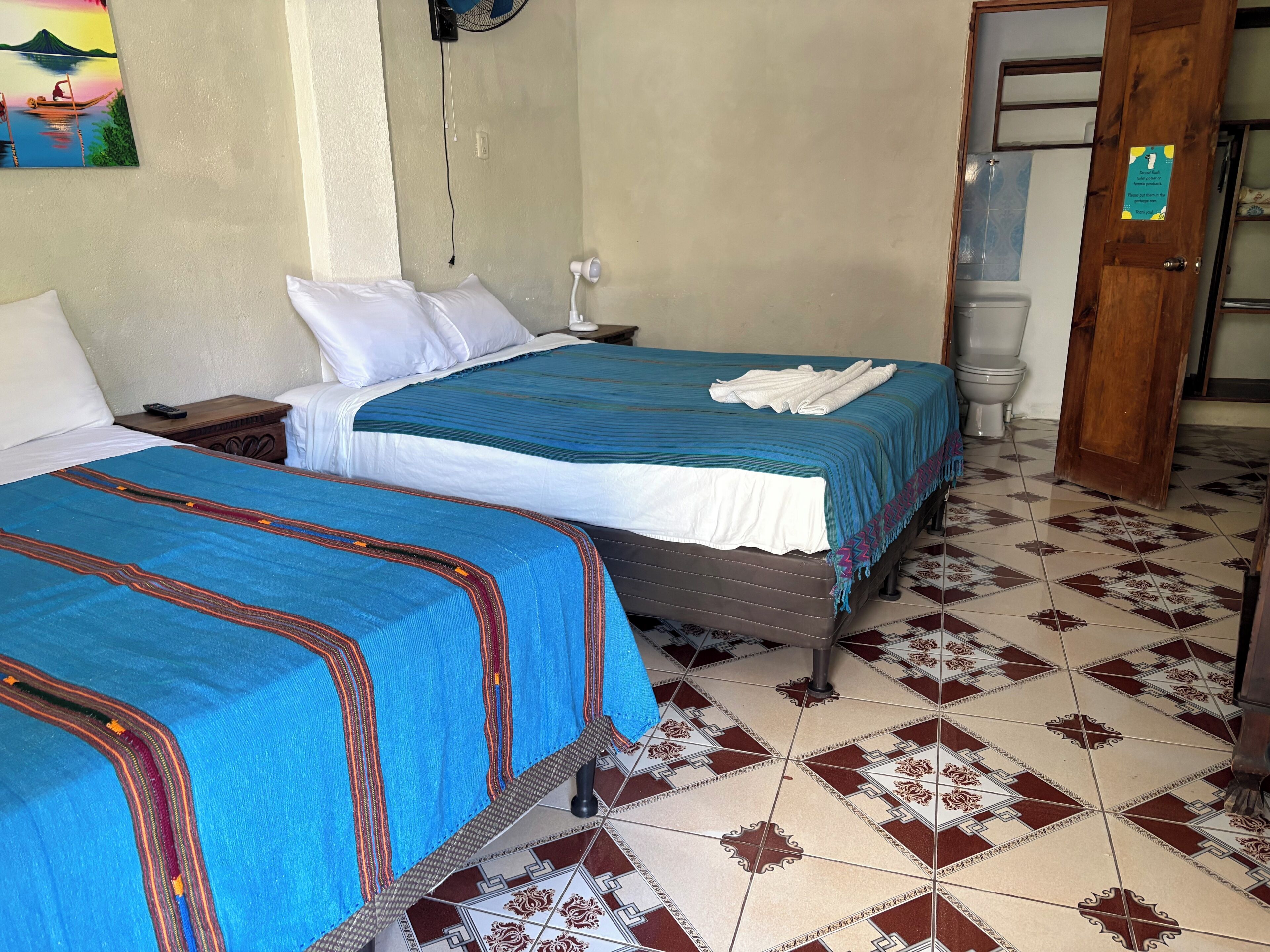 A hotel room with two beds, a nightstand, a wall-mounted fan, a toilet, and a door with a sign.