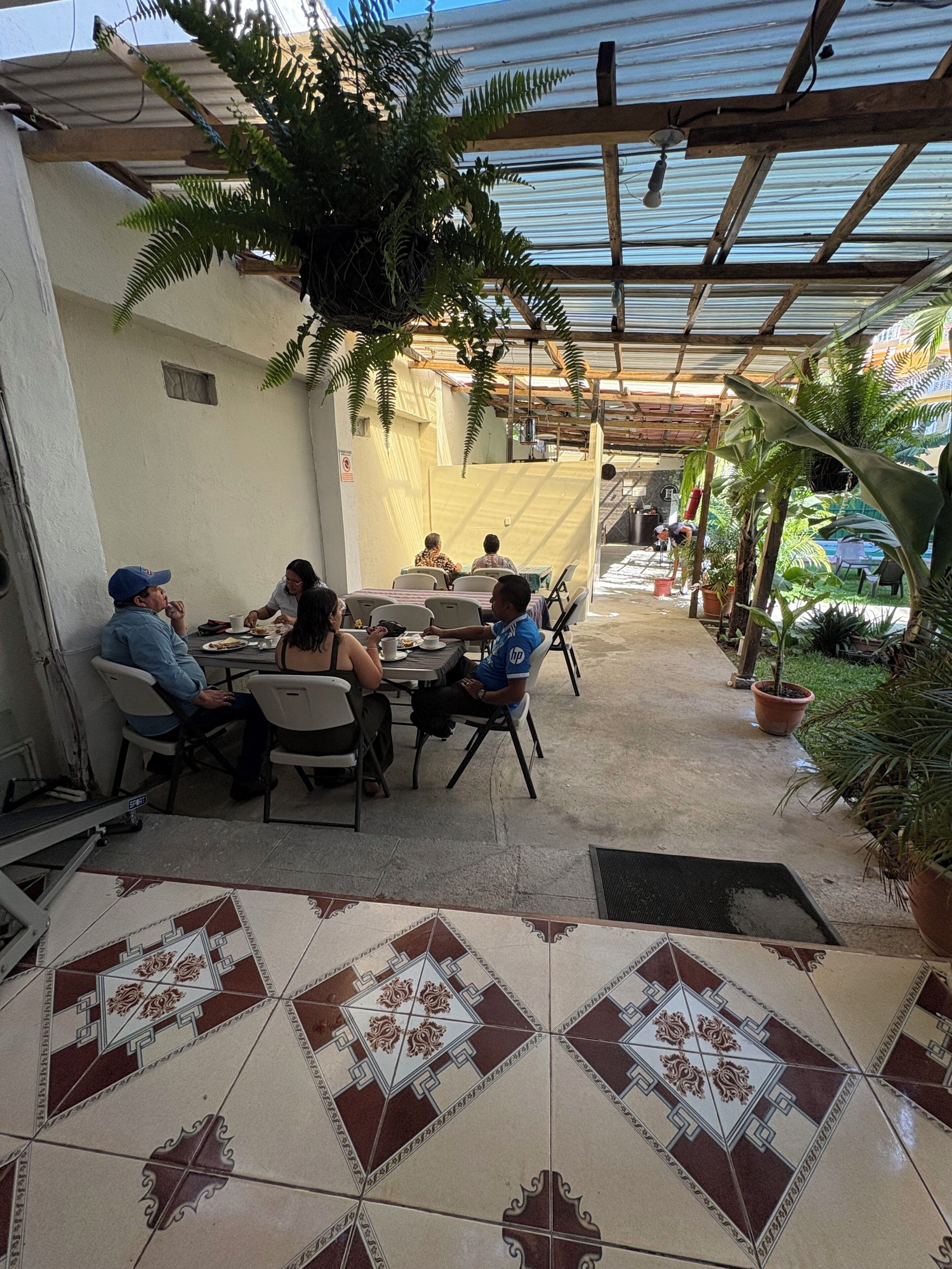 Une terrasse extérieure avec un sol carrelé, un groupe de personnes assises autour d’une table et des plantes en pot.