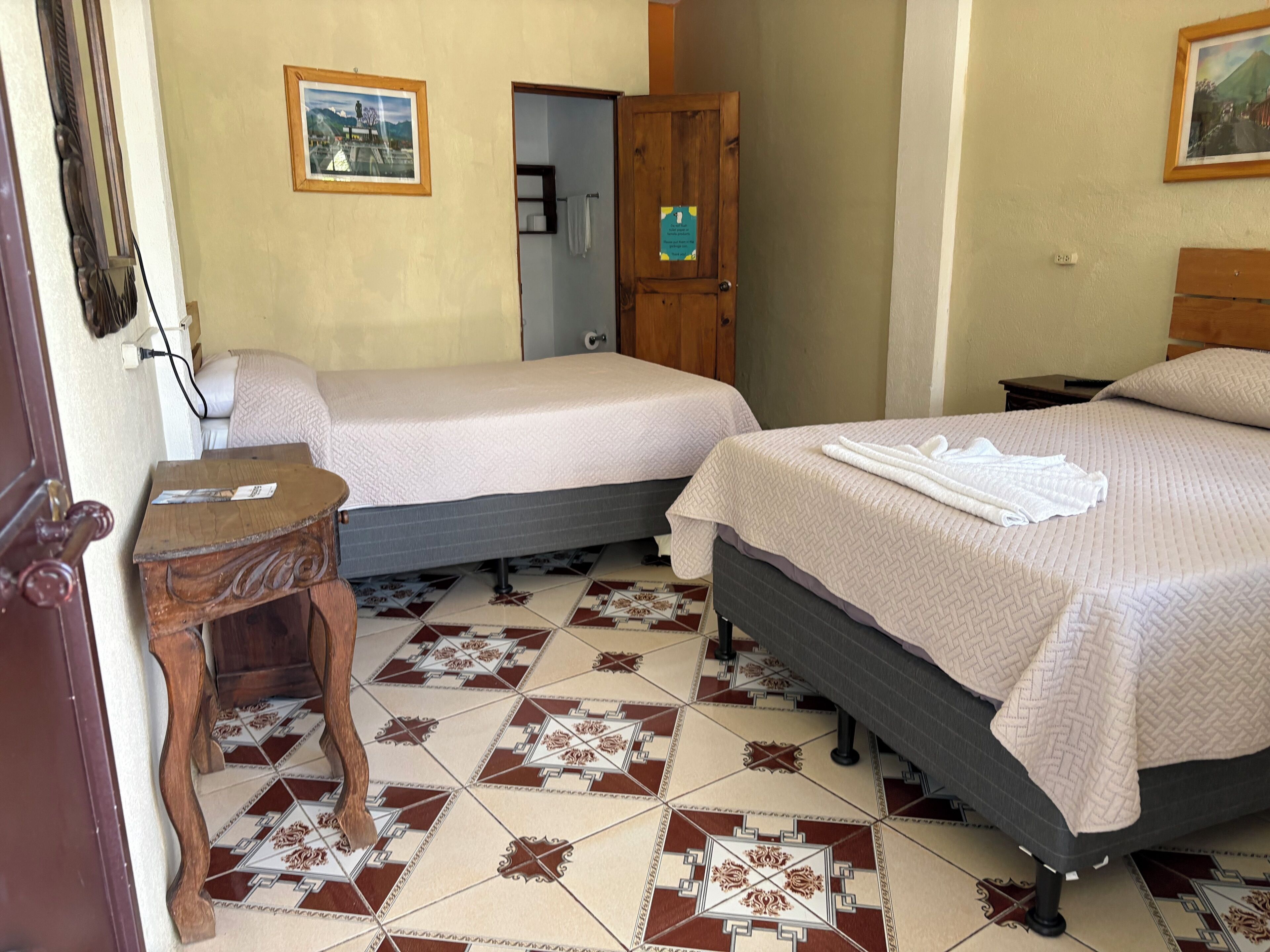Habitación de hotel con dos camas, una mesita de noche de madera, piso de baldosas y puerta que da a un baño.