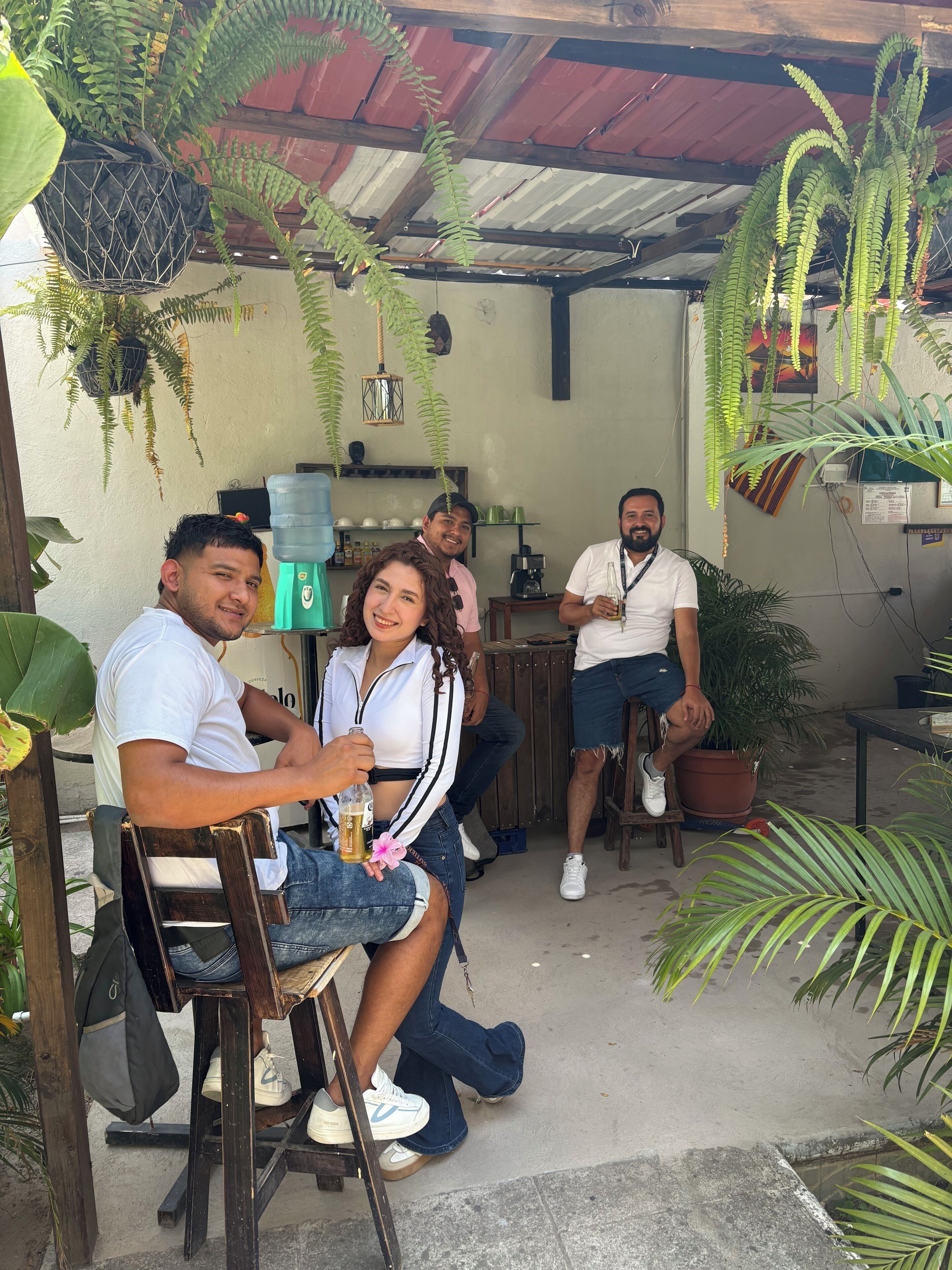 A group of people seated at a wooden table in a rustic setting with hanging plants and a coffee machine.