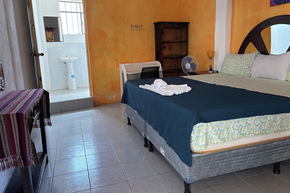 A hotel room with a bed, a bedside table, a chair, a wardrobe, a fan, and a bathroom visible through an open door.