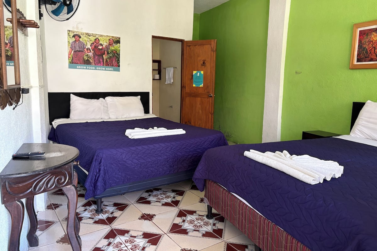 A hotel room with two beds, a desk with a remote control, a fan, and a green wall.