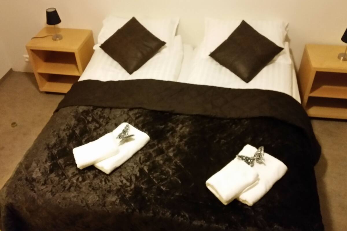 Een net opgemaakt bed met een zwart dekbed, twee witte handdoeken en twee zwarte kussens. Er staan twee houten nachtkastjes met lampen op elk.