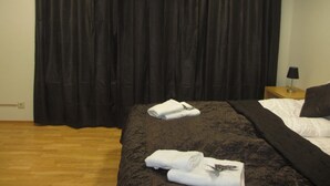 A hotel room with a large bed, dark curtains, a wooden floor, and a nightstand with a lamp.