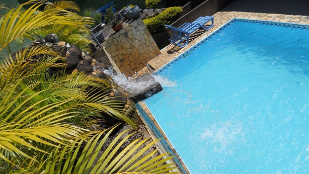 A swimming pool with a stone wall and water feature, surrounded by palm trees and potted plants. - Pousada O Meu Canto (Santa Rita de Jacutinga)