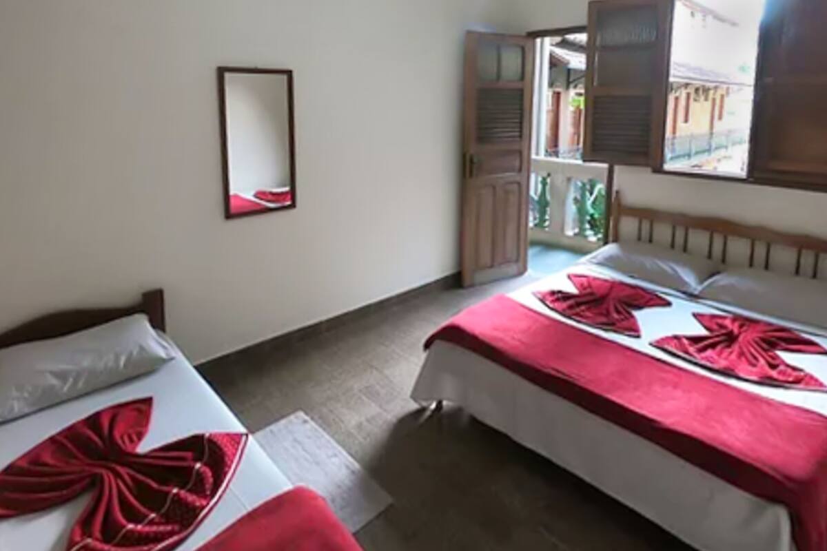 Quarto com duas camas, cada uma com um cobertor e travesseiro vermelhos. Há um espelho na parede e uma janela com persianas de madeira.