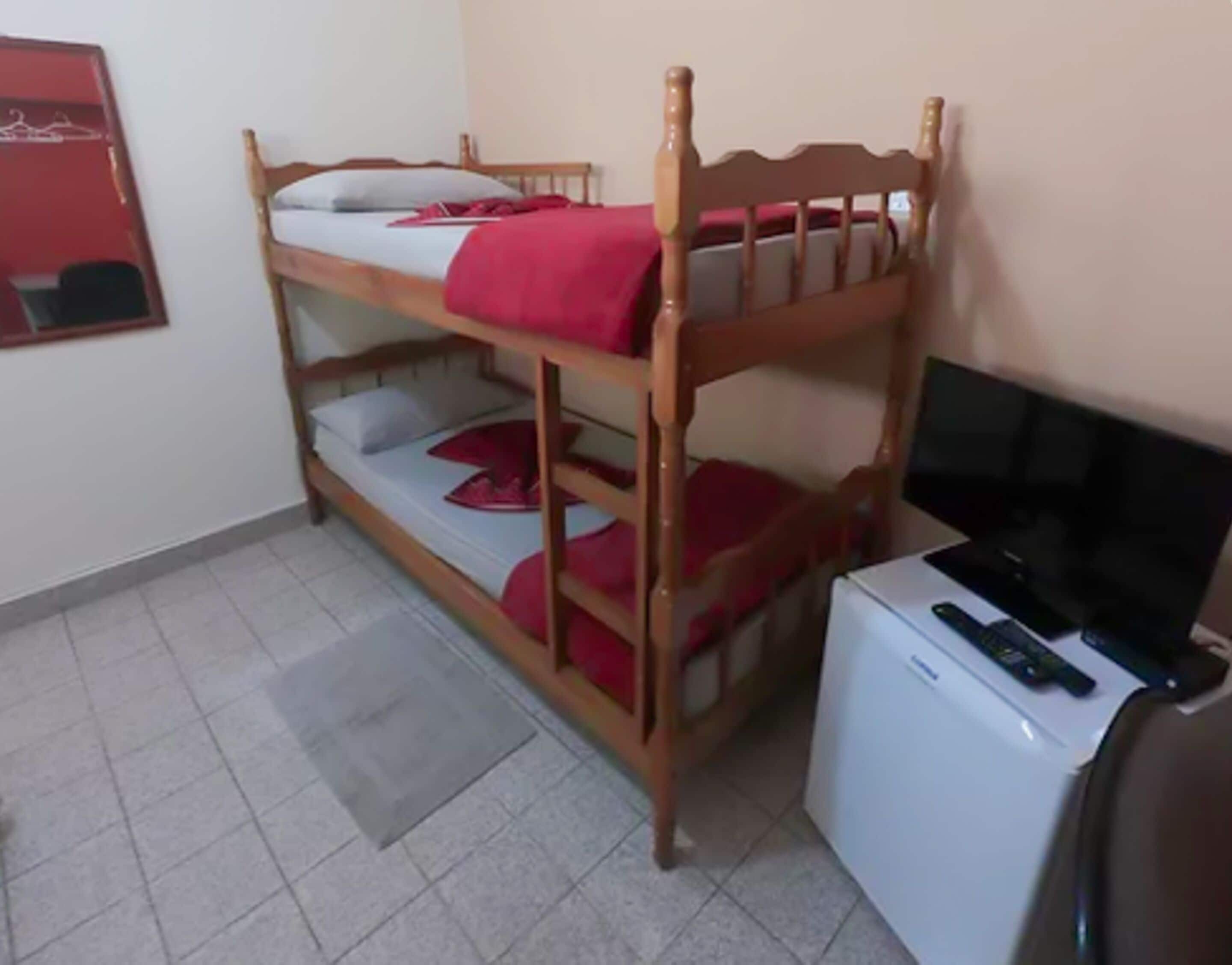 Uma cama beliche com um cobertor vermelho, um refrigerador branco pequeno e uma televisão em um suporte.