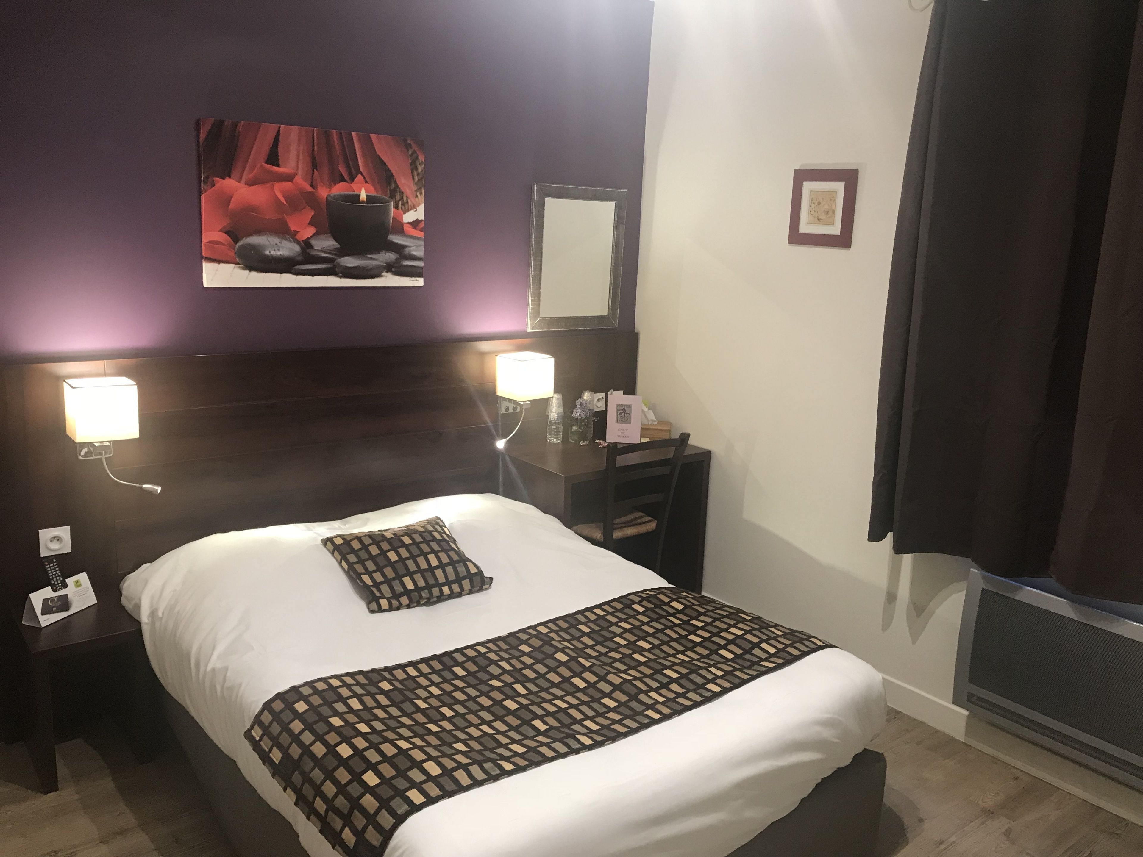 A hotel room with a bed, a nightstand, a lamp, a mirror, and a picture on the wall.