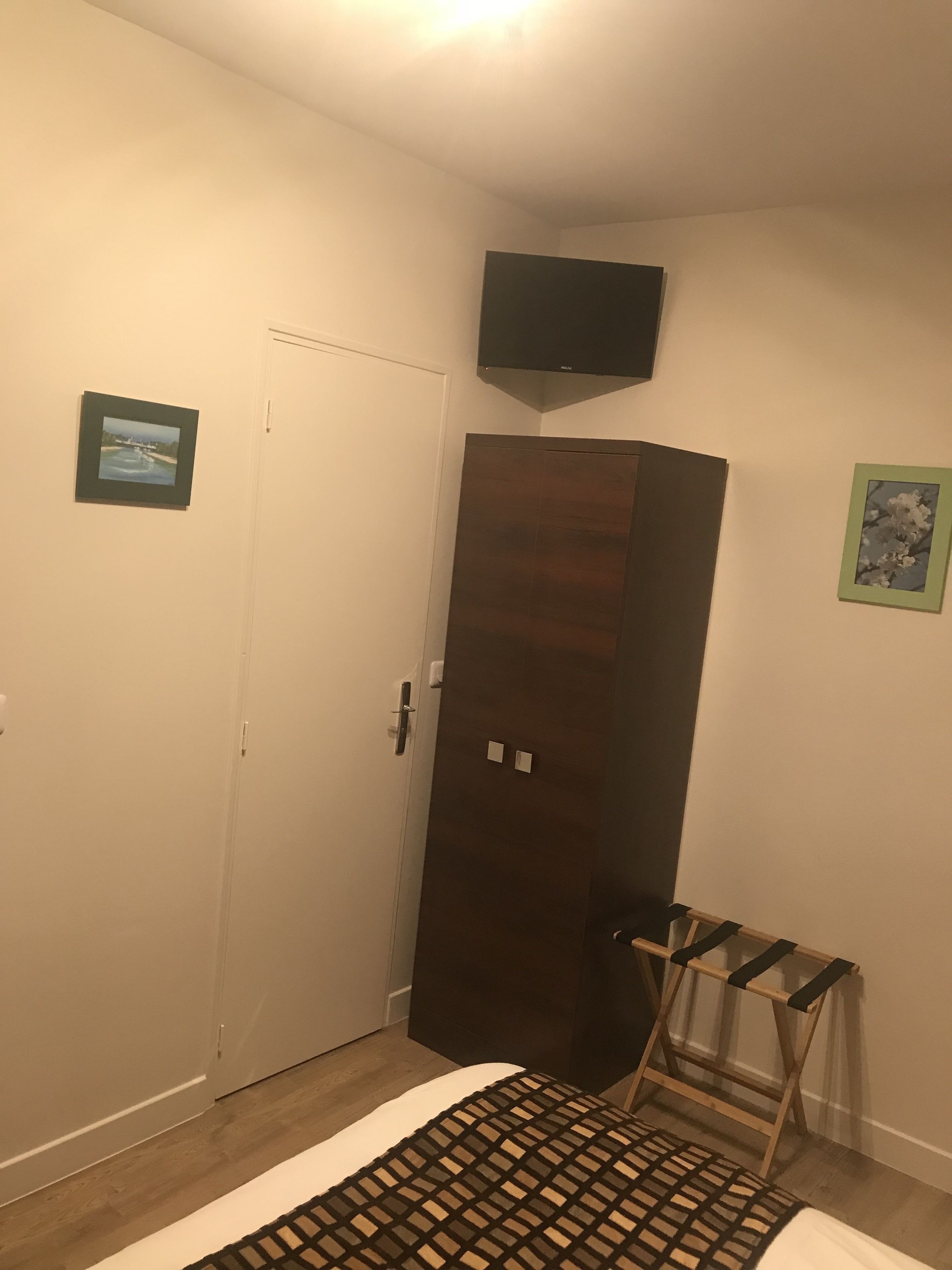 A room with a wooden wardrobe, a television mounted on the wall, and a small wooden rack.