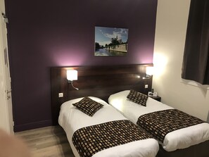 A hotel room with two beds, each with a patterned bedspread and matching pillows. There are wall-mounted lamps on either side of the beds, and a painting of a bridge is visible on the wall above the beds. - Hôtel les Iris (Les Andelys)