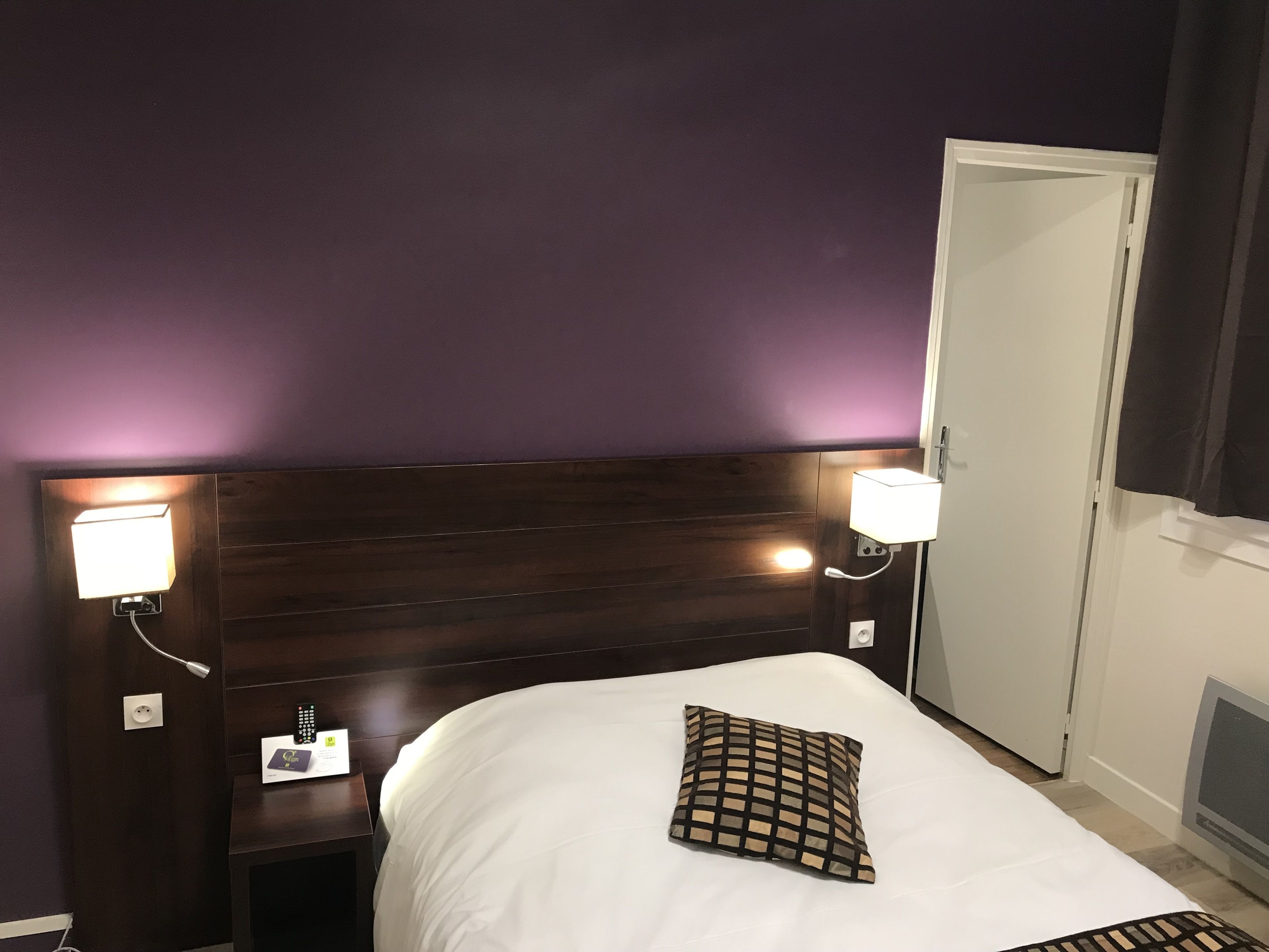 Habitación de hotel con tablero de cabecera de madera oscura, ropa de cama blanca y un cojín a cuadros, además de lámparas de noche.