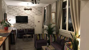 A living room with a brick wall, a flat-screen TV, a sofa set, a coffee table, and a potted plant. - Hôtel les Iris (Les Andelys)