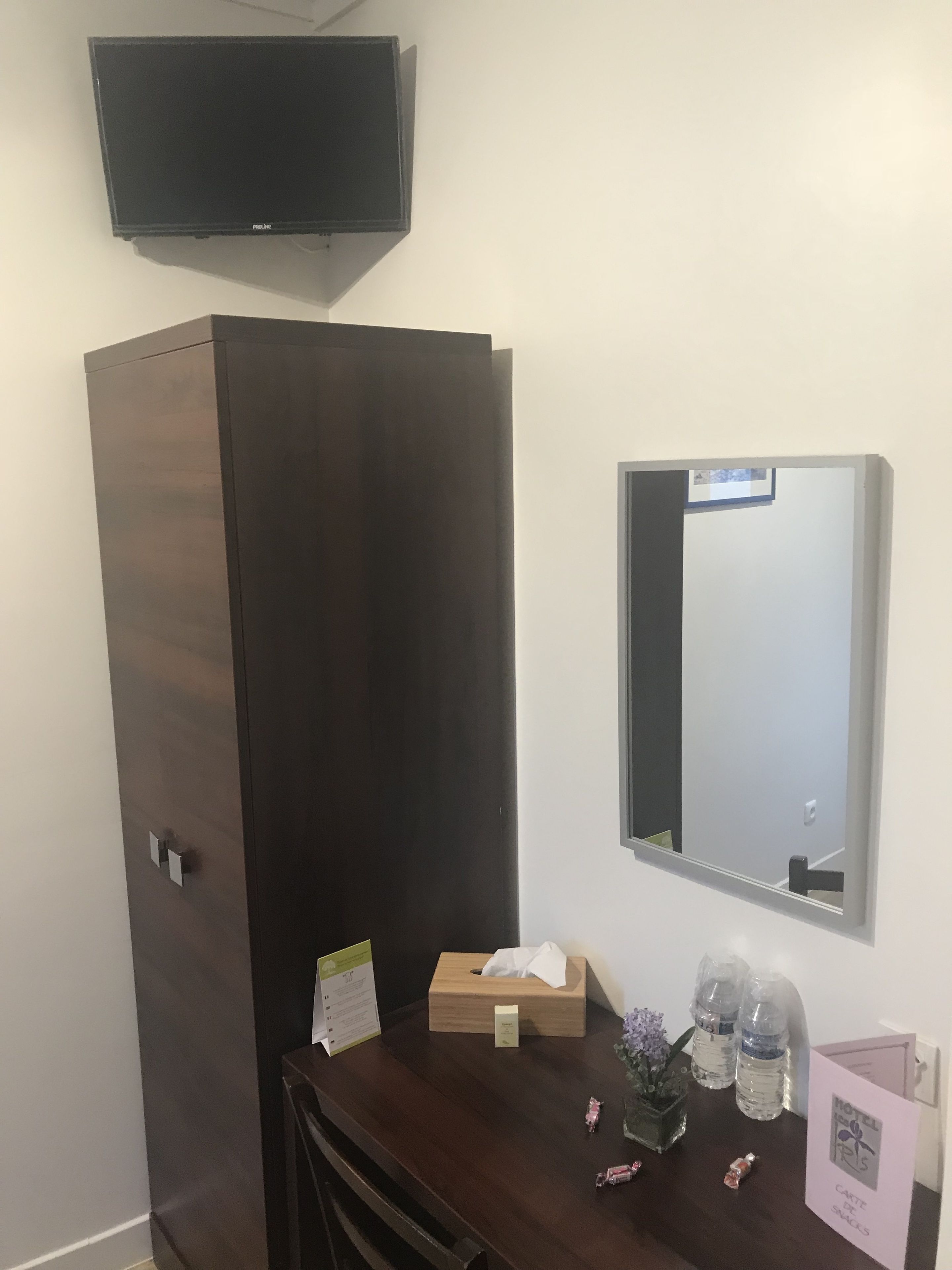A hotel room with a dark wooden wardrobe, a small wooden desk with a tissue box, a mirror, and a card stand with a hotel logo.