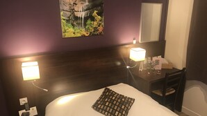 A hotel room with a bed, a nightstand, a desk with a lamp, a mirror, and a picture of a waterfall on the wall. - Hôtel les Iris (Les Andelys)