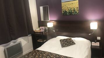 A hotel room with a bed, a desk, a chair, a mirror, and a wall-mounted light.