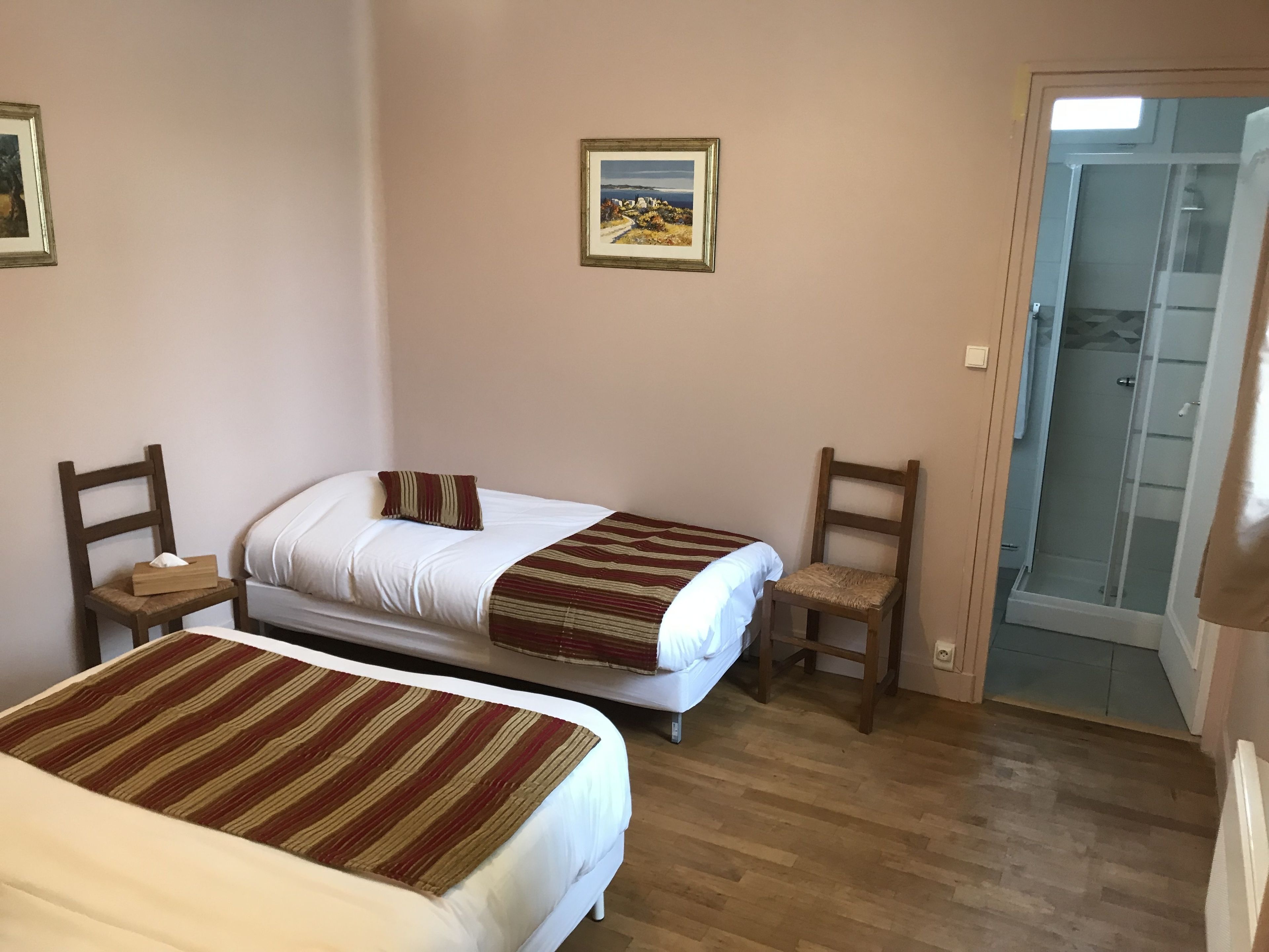 A room with two beds, a wooden floor, a door leading to a shower, and a framed picture on the wall.
