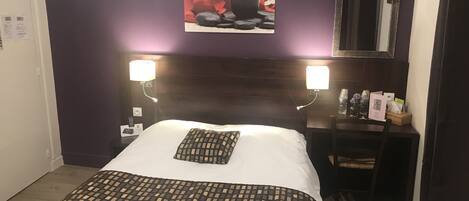 A hotel room with a bed, two bedside lamps, a desk with a chair, a mirror, and a wall-mounted picture.