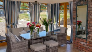 A sunlit room with wicker furniture, a glass table, and a view of a garden through large windows. - Castleview - Newton Steading (Kirriemuir)