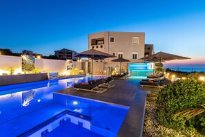 A modern outdoor pool area with lounge chairs and umbrellas, a well-lit pool, and a building in the background. - Talos Luxury Suites (Chania)
